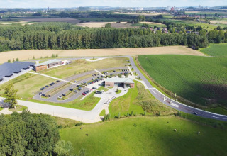 Luftfoto af glamping-overnatning med parkeringsplads, grønne marker og skov i baggrunden, solrig dag