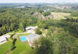 Luftfoto af glamping-område med swimmingpool, hytter i skov og udsigt til sø og grønne marker.