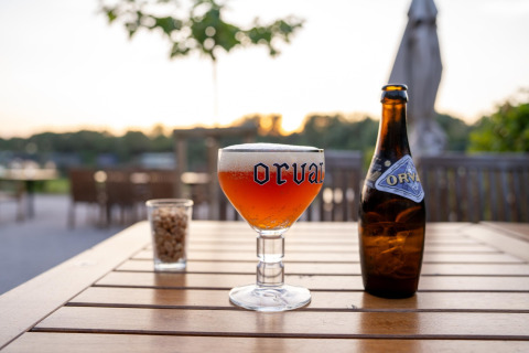 Un vaso de cerveza Orval y una botella sobre una mesa de madera al aire libre al atardecer en glamping.
