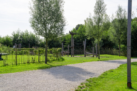 Grüner Außenbereich einer Glamping-Unterkunft mit Holzspielplatz, Bäumen und Kiesweg im Sonnenschein.