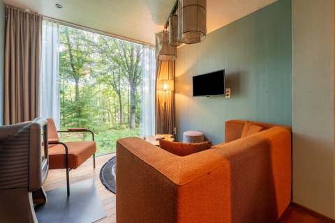 Sala de glamping moderna con grandes ventanales, sofá naranja y vistas al bosque y la naturaleza.