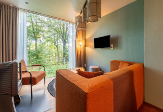Sala de glamping moderna con grandes ventanales, sofá naranja y vistas al bosque y la naturaleza.