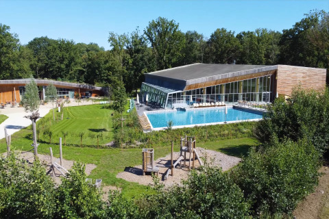 Modern tree house resort in Belgium featuring a large outdoor pool, green lawns, and children’s playground.