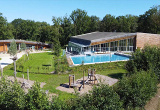 Modern tree house resort in Belgium featuring a large outdoor pool, green lawns, and children’s playground.