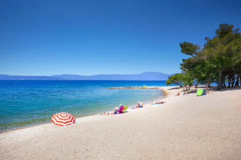 Strand bij Aminess Gaia Green Villas Njivice in Kroatië met zonaanbidders, blauwe zee en bomen.