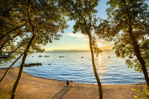 Due persone sedute su una spiaggia al tramonto guardano il mare e le barche, circondate da alberi in Croazia.