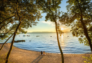Due persone sedute su una spiaggia al tramonto guardano il mare e le barche, circondate da alberi in Croazia.