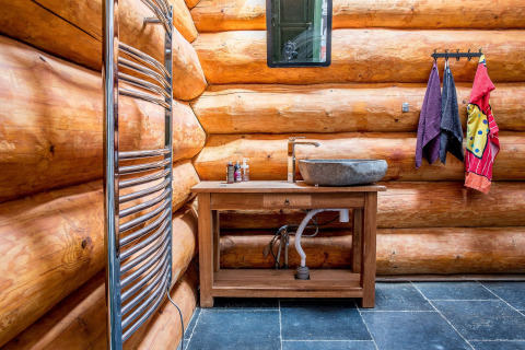 Bagno rustico in cabina di tronchi con lavabo in pietra, specchio, asciugamani e tubature a vista.