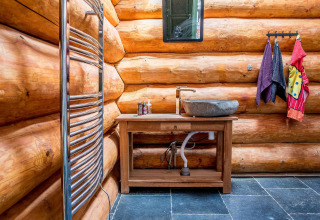 Bagno rustico in cabina di tronchi con lavabo in pietra, specchio, asciugamani e tubature a vista.