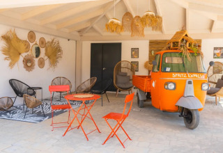 Outdoor lounge with bright orange Spritz Express tuk-tuk, eclectic decor, and modern garden chairs in Italy.