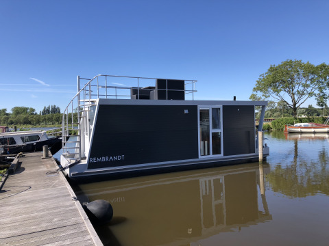 Moderne woonboot genaamd Rembrandt aangemeerd aan een steiger bij Marina Parcs Amsterdam onder een blauwe lucht.