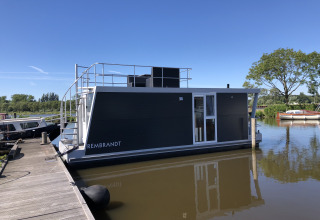 Moderne woonboot genaamd Rembrandt aangemeerd aan een steiger bij Marina Parcs Amsterdam onder een blauwe lucht.