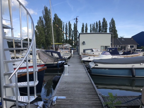 Een houten steiger met aangemeerde boten bij Marina Parcs Amsterdam, met moderne gebouwen op de achtergrond.