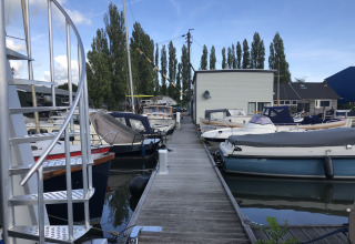 Een houten steiger met aangemeerde boten bij Marina Parcs Amsterdam, met moderne gebouwen op de achtergrond.