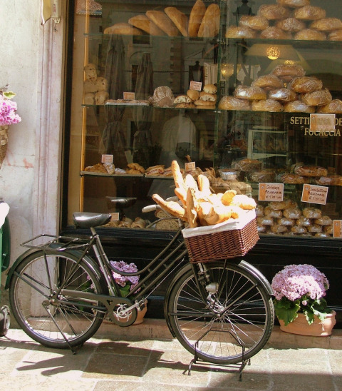 Een fiets met een mand vol stokbroden staat voor een etalage van een bakkerij met verschillende soorten brood.