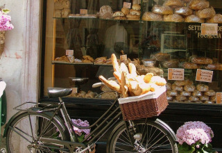 Een fiets met een mand vol stokbroden staat voor een etalage van een bakkerij met verschillende soorten brood.