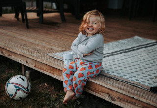 Lachend kind zit op houten terras bij Camping Emmen in Drenthe, Nederland, met bal en mok op de grond.