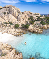 Bahía rocosa con agua cristalina y una persona nadando, en un parque de glamping visto desde arriba.
