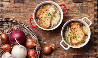 Dos cuencos de sopa de cebolla francesa con pan y queso, rodeados de cebollas y ajo sobre mesa de madera.