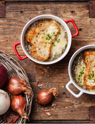 Dos cuencos de sopa de cebolla francesa con pan y queso, rodeados de cebollas y ajo sobre mesa de madera.