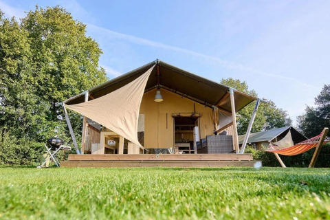 Tenda safari Country con terrazza in legno, barbecue e amaca su un prato verde sotto cielo azzurro.