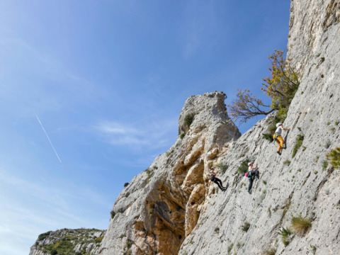 Three climbers scaling a steep rock face beneath a blue sky at La Vallée Heureuse campsite in Provence, France.