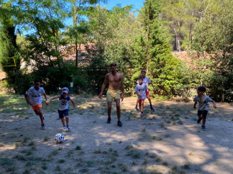 Children and an adult playing soccer on a dirt field surrounded by trees at La Vallée Heureuse campsite, France.