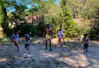 Bambini e un adulto giocano a calcio su un campo sterrato circondato da alberi al campeggio La Vallée Heureuse.