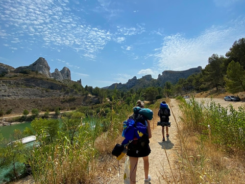 Escursionisti con zaini che camminano su un sentiero soleggiato vicino a montagne e un fiume in Provenza-Alpi-Costa Azzurra.