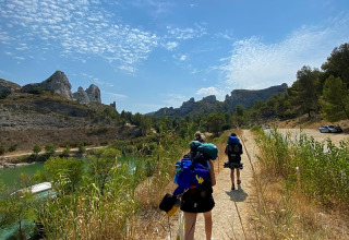 Hikers with backpacks walking on a sunny trail by mountains and a river in Provence-Alpes-Côte d’Azur, France.