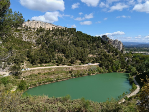View of a green pond surrounded by forested hills near Orgon in Provence-Alpes-Côte d’Azur, France, under blue sky.