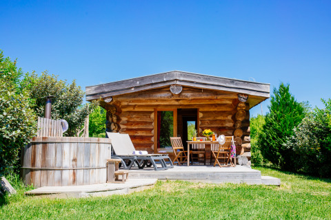 Blockhütte Trappeur mit Spa, Terrasse, Liegestühlen und Whirlpool in grüner Natur unter blauem Himmel.