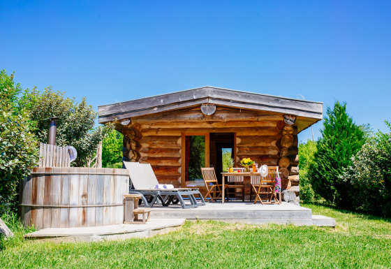 Blockhütte Trappeur mit Spa, Terrasse, Liegestühlen und Whirlpool in grüner Natur unter blauem Himmel.