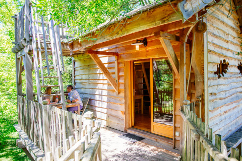 Et billede af et hyggeligt træhus kaldet Treehouse Palombière med to personer, der sidder på terrassen.