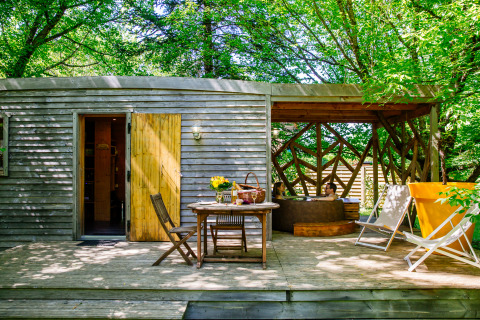 Outdoor wooden deck at a lodge with dining table, loungers, and two people relaxing in a hot tub beneath trees.