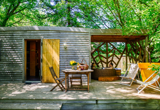 Outdoor wooden deck at a lodge with dining table, loungers, and two people relaxing in a hot tub beneath trees.