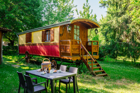 Cabina de carro gitano colorido en jardín verde, con mesa de desayuno al aire libre rodeada de árboles.