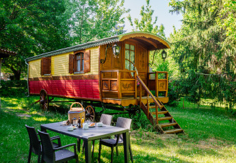 Cabina de carro gitano colorido en jardín verde, con mesa de desayuno al aire libre rodeada de árboles.
