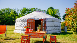 Traditionelle Jurte mit bunten Möbeln und Früchten auf einem sonnigen Feld, umgeben von Bäumen.