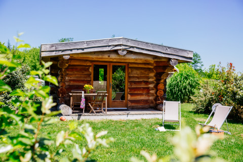 Log cabin Trappeur nature house with green lawn, deck chairs, and patio under a clear blue sky.