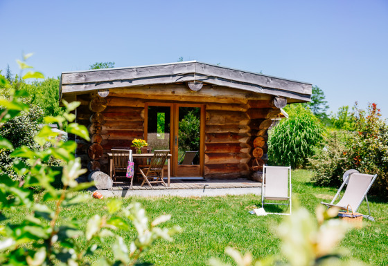 Cabane en bois Trappeur entourée de nature, pelouse verdoyante, chaises longues et meubles d’extérieur.
