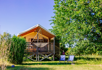 Safari tent op verhoog met hangmat en twee ligstoelen op het gras onder een grote, groene boom.