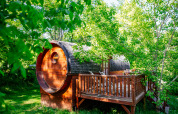 A round wooden cabin with a deck surrounded by lush green trees and sunlight in a forested area.
