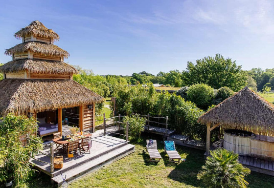 Indonesisch geïnspireerd natuurhuisje met spa, rieten dak, houten terras en ligstoelen in groene tuin.