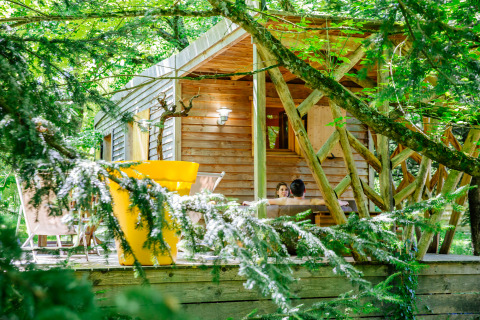 Cozy wooden cabin in the forest with two people relaxing on the terrace, surrounded by lush trees.
