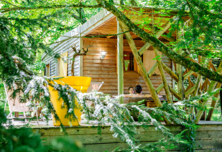 Cozy wooden cabin in the forest with two people relaxing on the terrace, surrounded by lush trees.