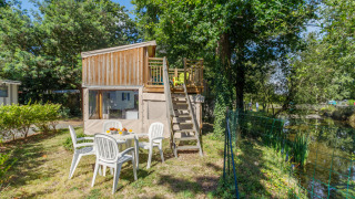 Tente Campétoile avec terrasse en bois et table de jardin près d’un étang entouré d’arbres verts.