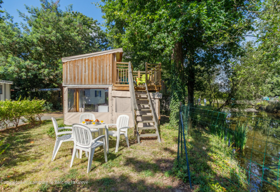 Tienda Campétoile con terraza de madera, mesa de exterior, junto a un estanque y rodeada de árboles.