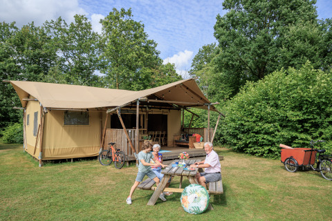 Famiglia al tavolo da picnic davanti alla tenda safari «Vogelkers» al parco Kaps nei Paesi Bassi, con biciclette.