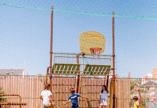 Children play football under a basketball hoop on an outdoor court at Flower Camping La Pointe du Talud.